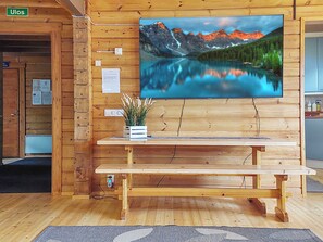 Interior de madera con un banco de madera, un televisor empotrado en la pared mostrando un lago de montaña, y una pequeña planta sobre el banco.