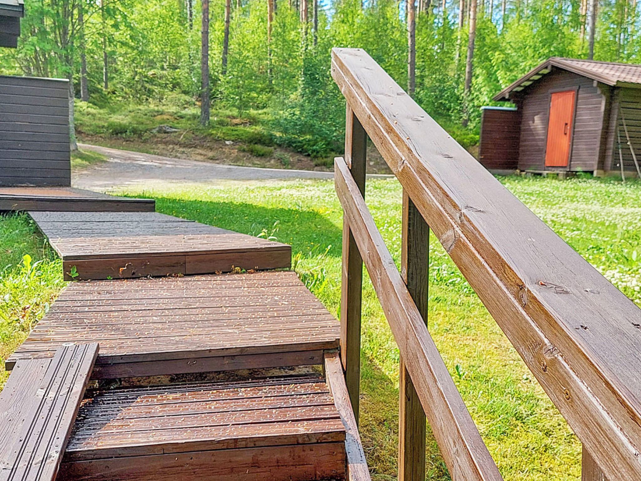Une terrasse en bois mène à un bâtiment doté d’une porte rouge, qui est entouré de verdure et bordé par une zone boisée.