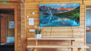 A wooden interior with a wooden bench, a wall-mounted TV displaying a mountain lake, and a small plant on the bench.