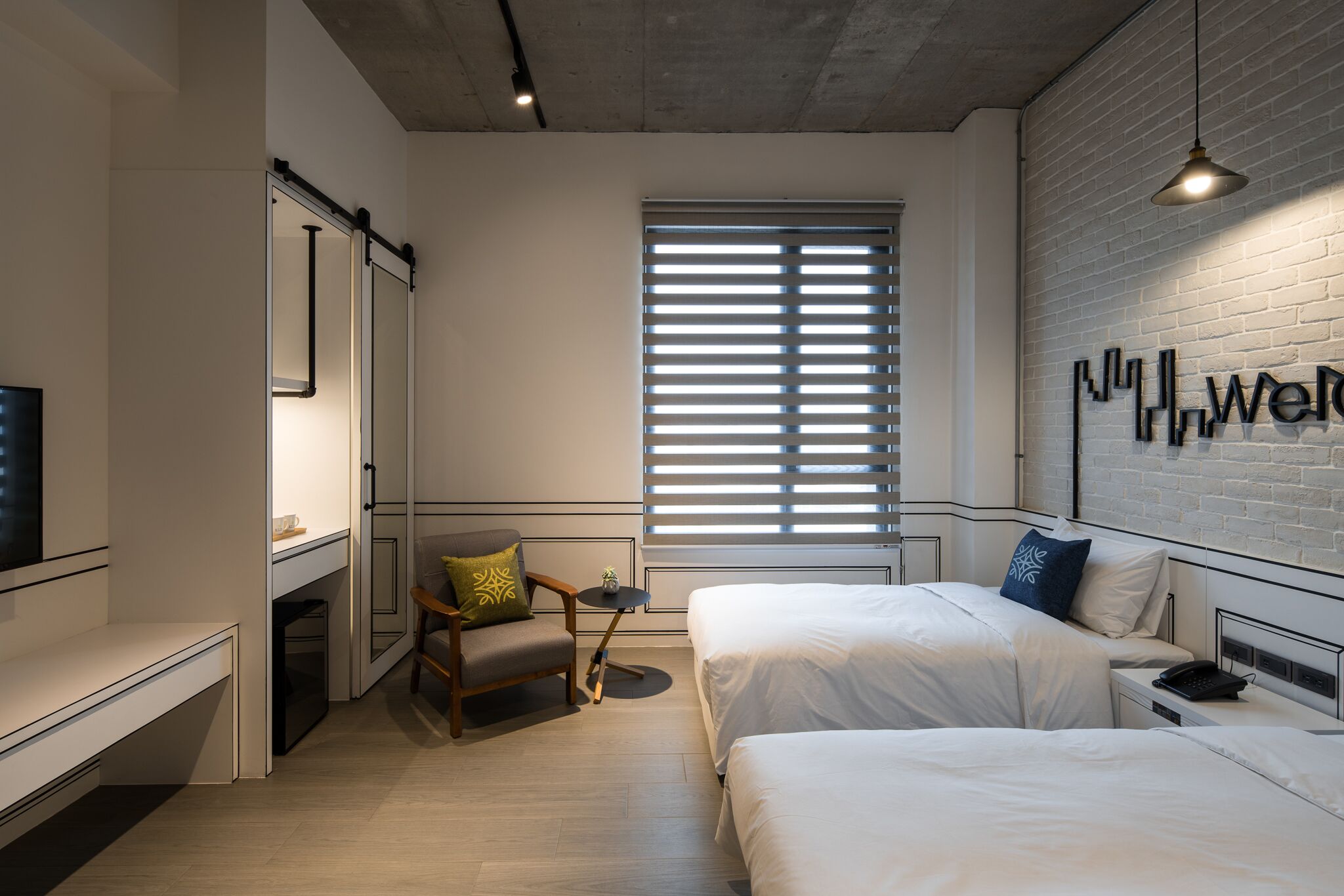 A modern hotel room with two beds, a small seating area, a TV, and a large window with blinds.