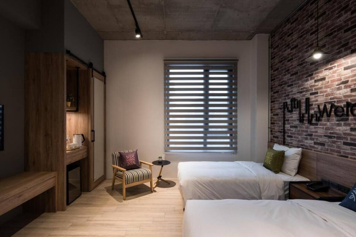 Une chambre d’hôtel avec deux lits, une petite table, une chaise, une fenêtre avec des stores et un mur en briques sur lequel est affiché un panneau.