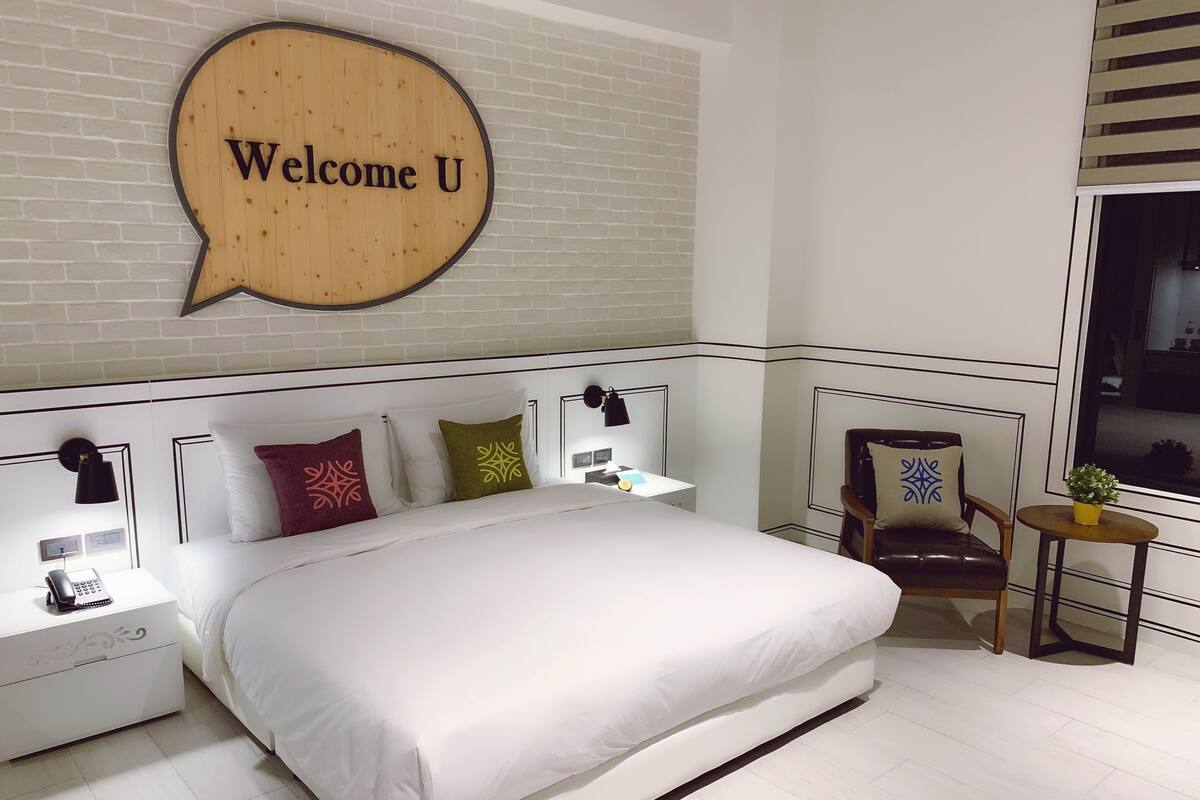 Une chambre d’hôtel moderne avec un grand lit, un petit bureau, une chaise et une fenêtre avec des stores.