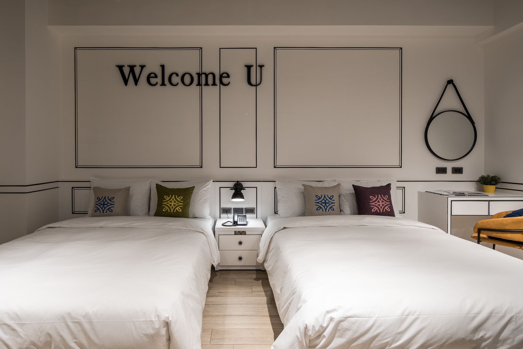 A modern hotel room with two beds, a bedside table with a lamp, a small desk, and a wall-mounted circular decoration.