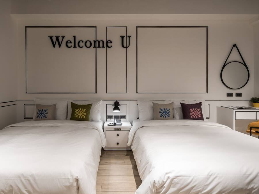 A modern hotel room with two beds, a bedside table with a lamp, a small desk, and a wall-mounted circular decoration.
