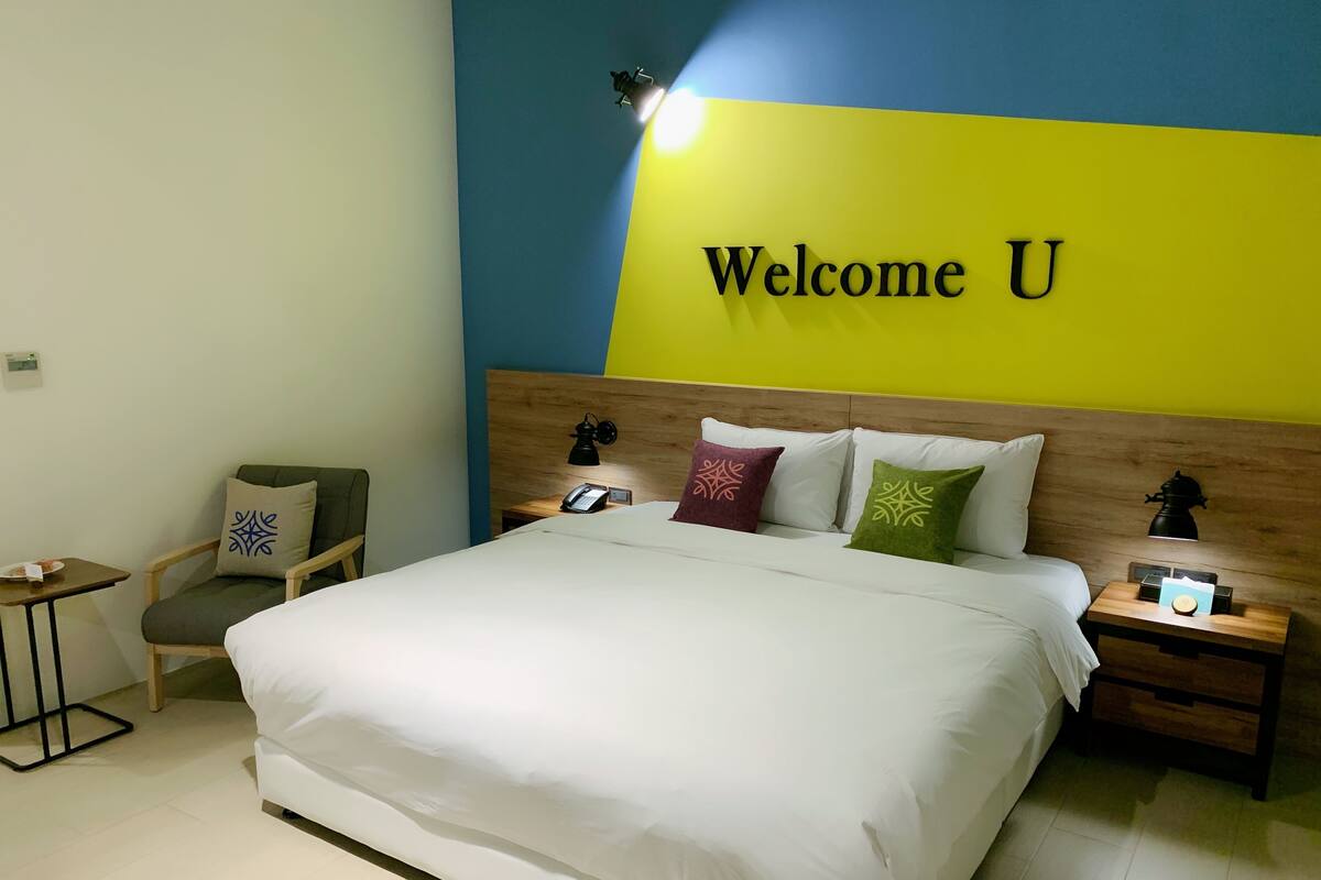 Une chambre d’hôtel avec un lit, une chaise, une table de chevet avec une lampe et un mur orné d’une inscription « Bienvenue ».