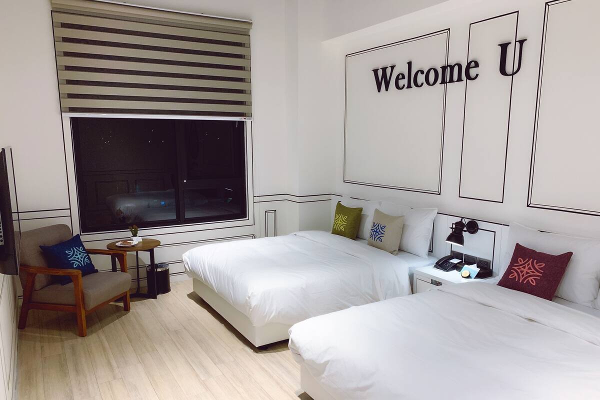 Une chambre d’hôtel avec deux lits, un bureau, une chaise, une fenêtre avec des stores et une pancarte « Bienvenue » accrochée au mur.