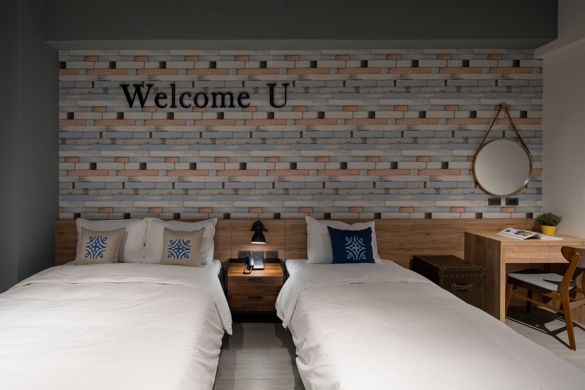 Habitación de hotel con dos camas, un escritorio de madera, una mesita pequeña, una lámpara y una pared con un cartel que dice 'Welcome U'.