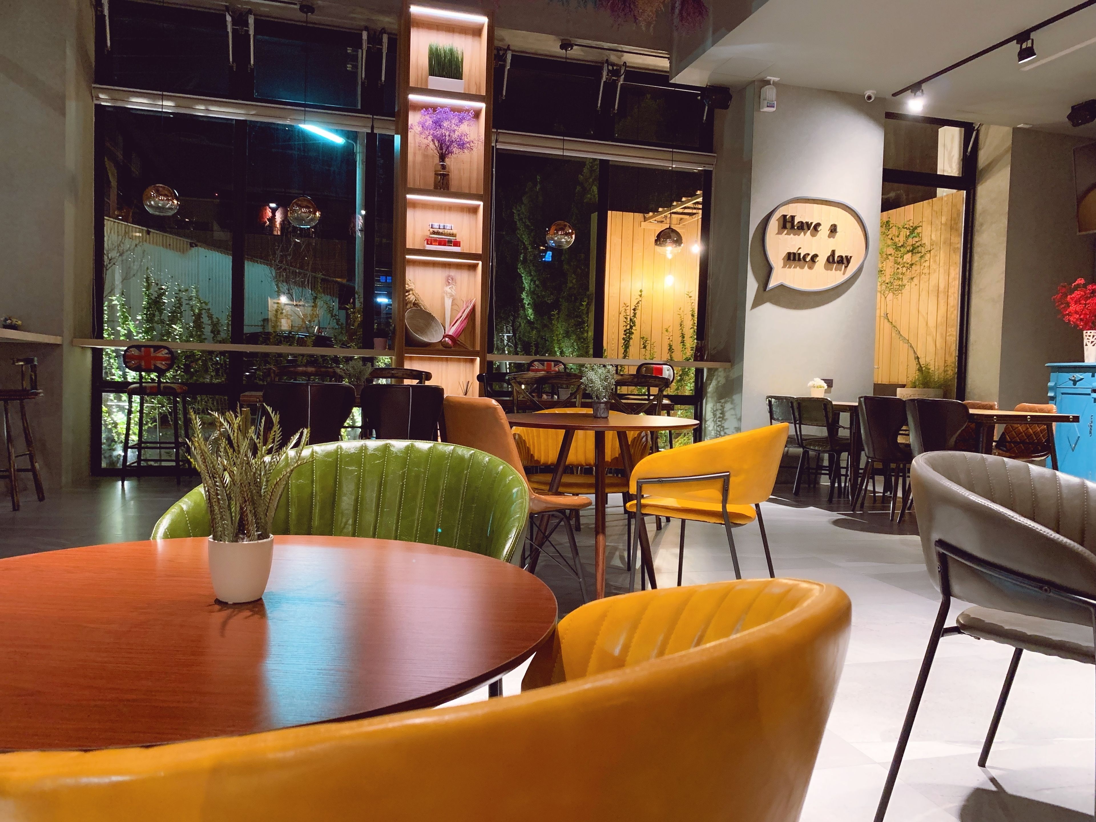 Un café moderno con mesas y sillas de madera, un amplio ventanal con plantas y un letrero que dice "Have a nice day".