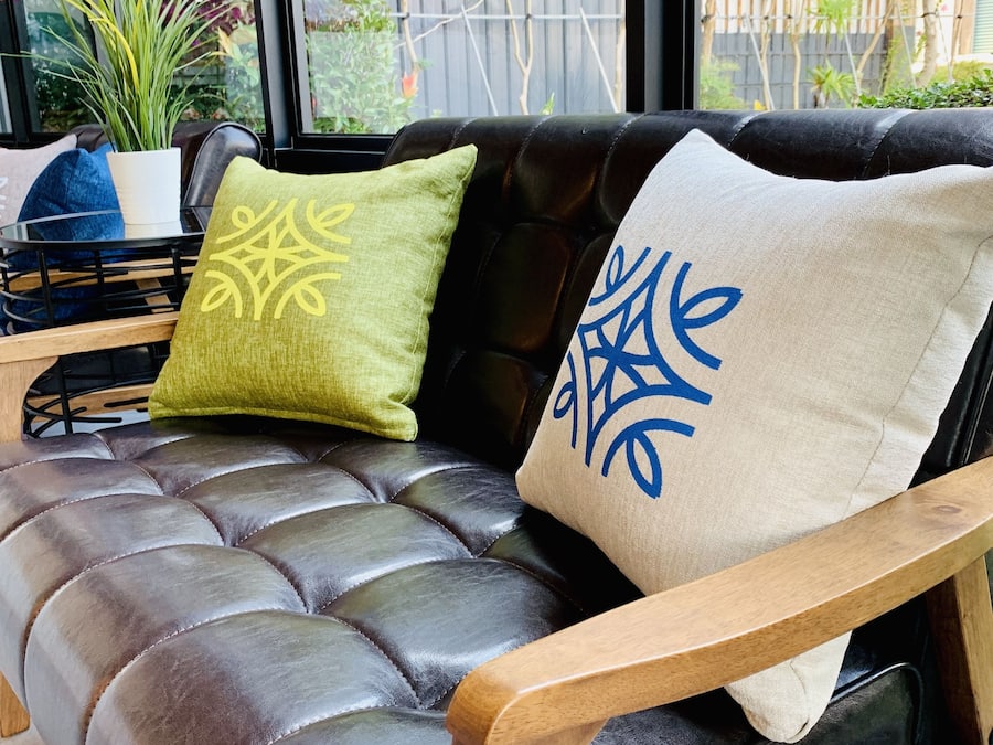 A leather sofa with wooden armrests, two cushions with embroidered designs, and a potted plant on a glass table beside it.