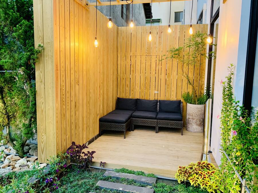 Una terraza de madera con un sofá negro, bombillas colgantes y plantas en macetas.