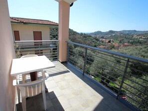 A balcony with a table and chairs, a view of a hilly landscape, and a building with a tiled roof in the background.