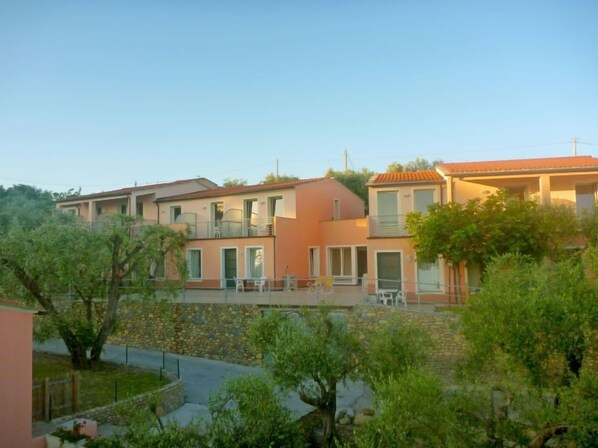 A row of modern, two-story buildings with balconies, surrounded by greenery and a stone wall.