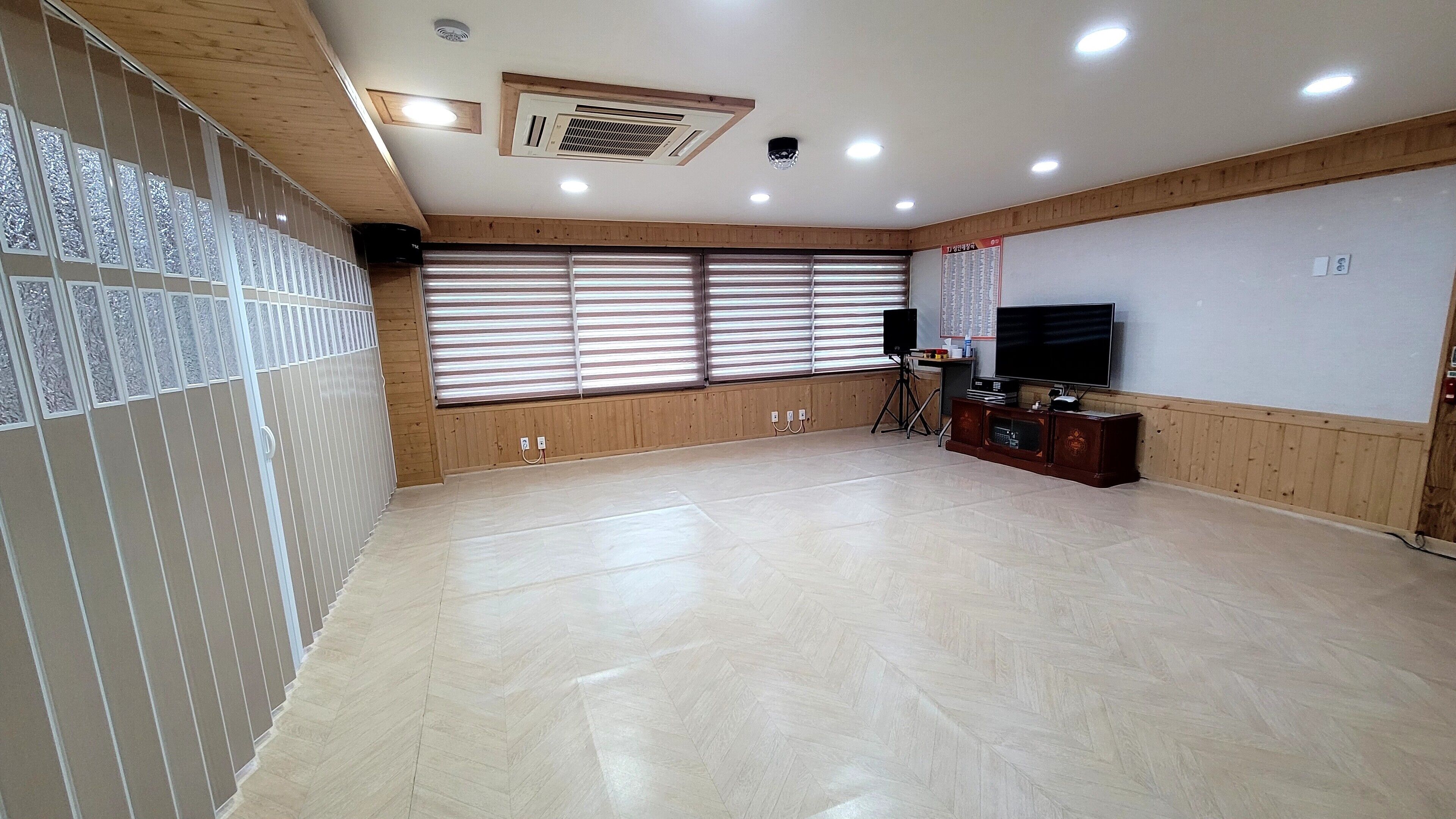 A spacious room with a large window, wooden paneling, a flat-screen TV, and a herringbone-patterned floor.