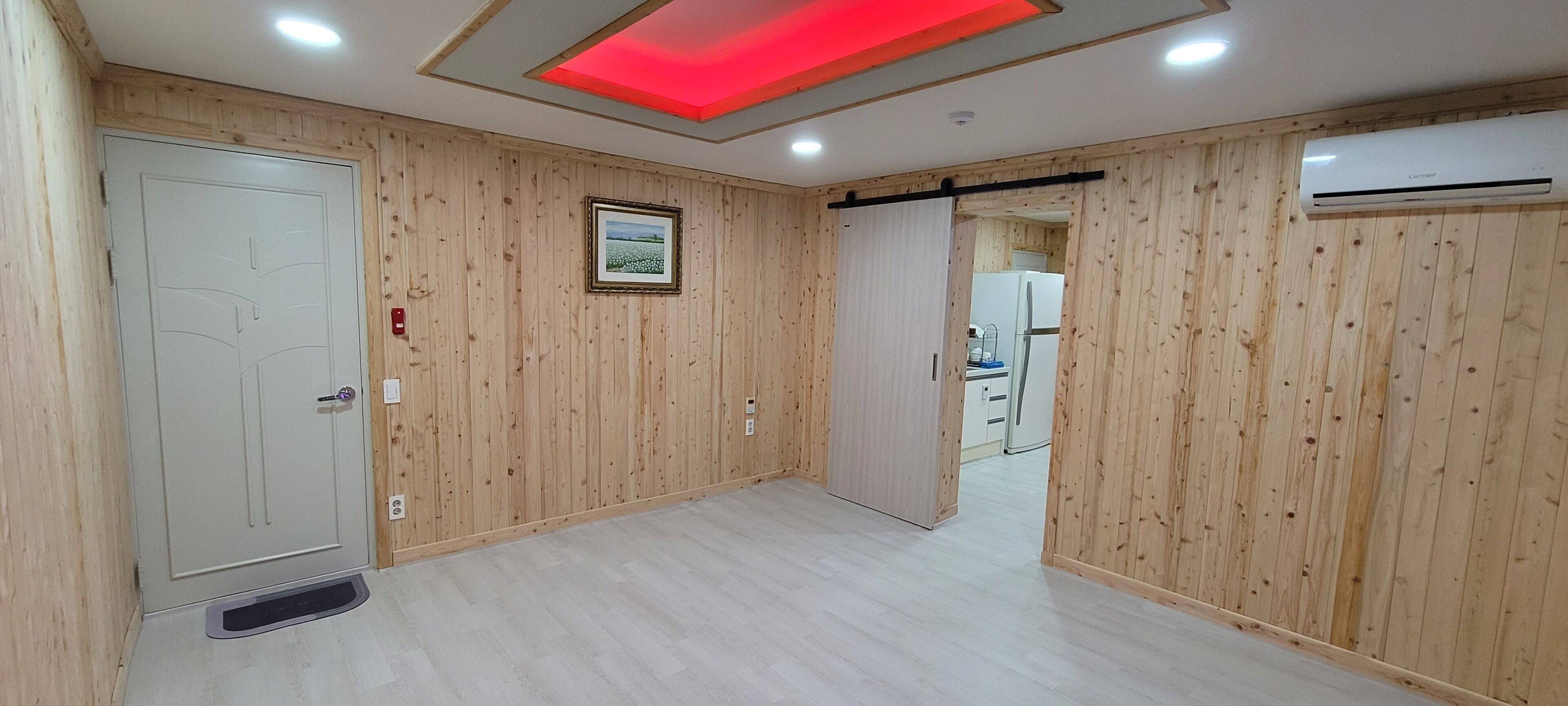 A room with wooden paneling, a white door, a framed picture on the wall, and a visible kitchen area with a refrigerator.