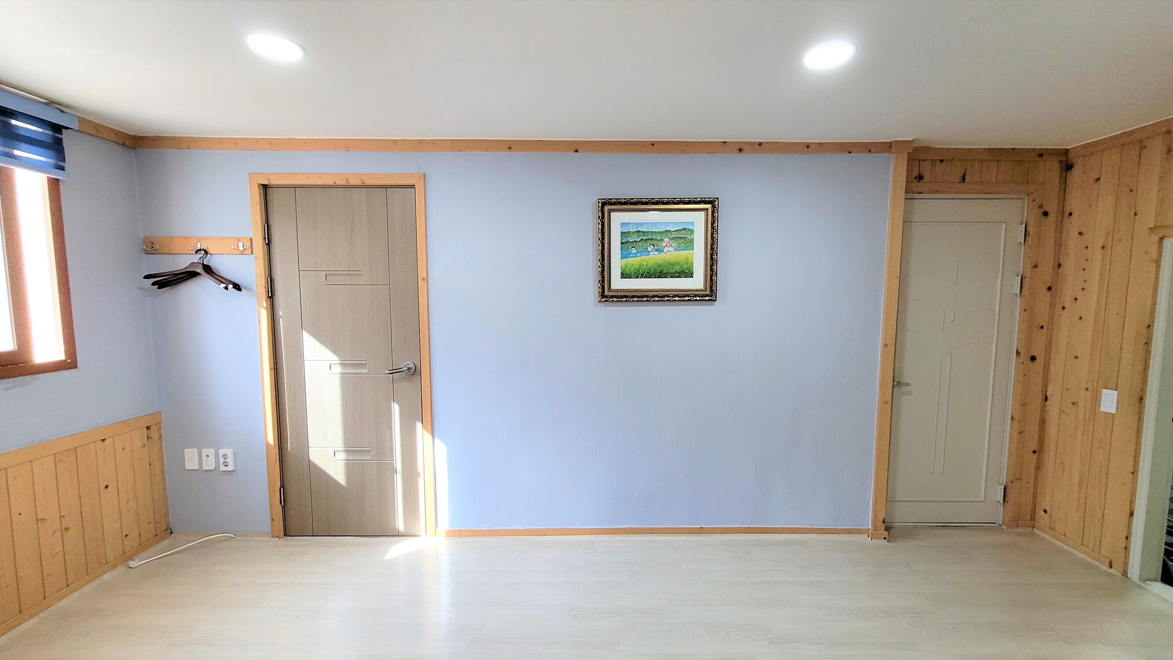 A room with light blue walls, wooden paneling on the lower half, a door with a handle, a framed picture on the wall, and a window with blinds.