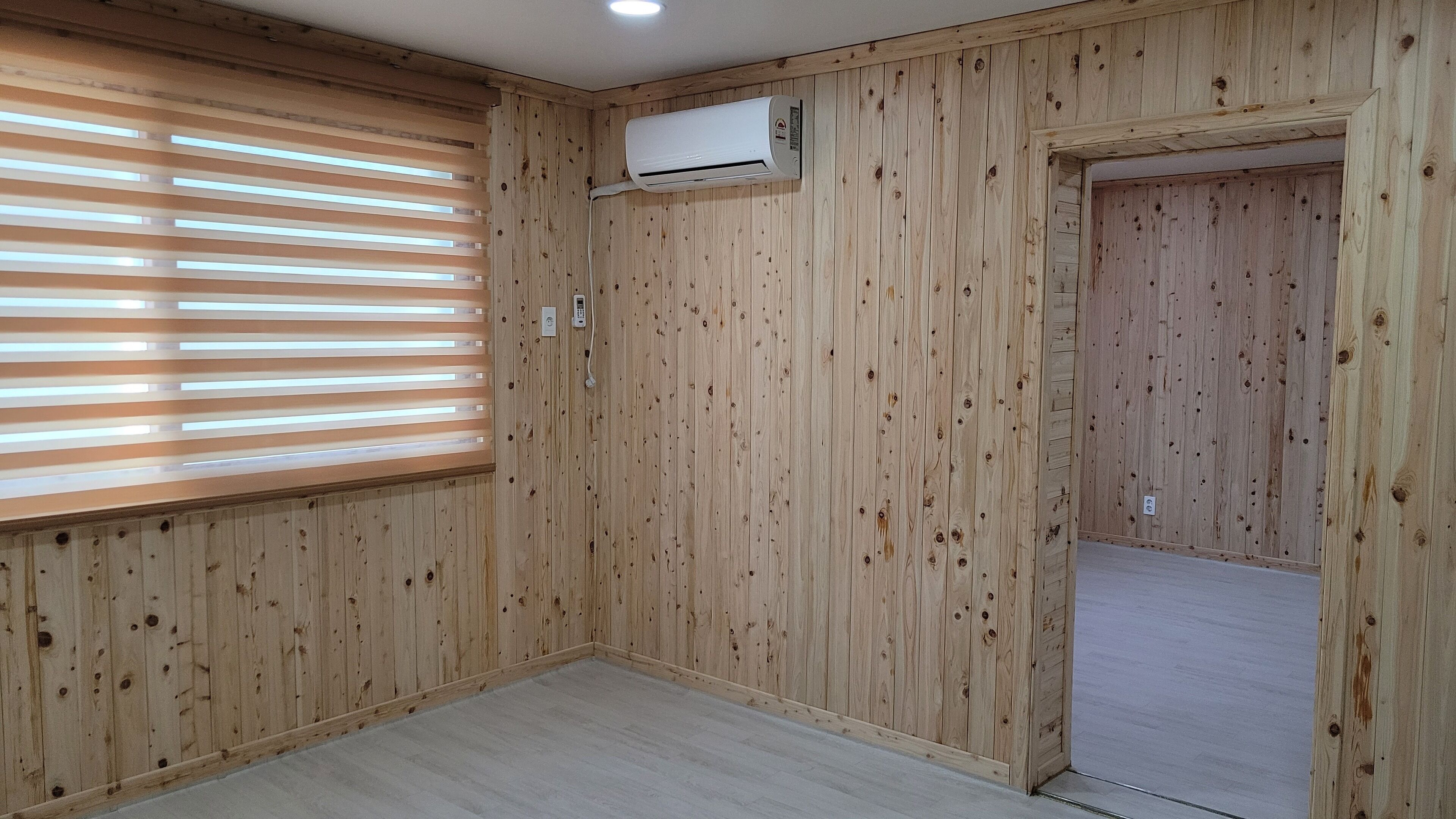 Una habitación con paredes de paneles de madera, un equipo de aire acondicionado instalado en el techo y una ventana con persianas horizontales.