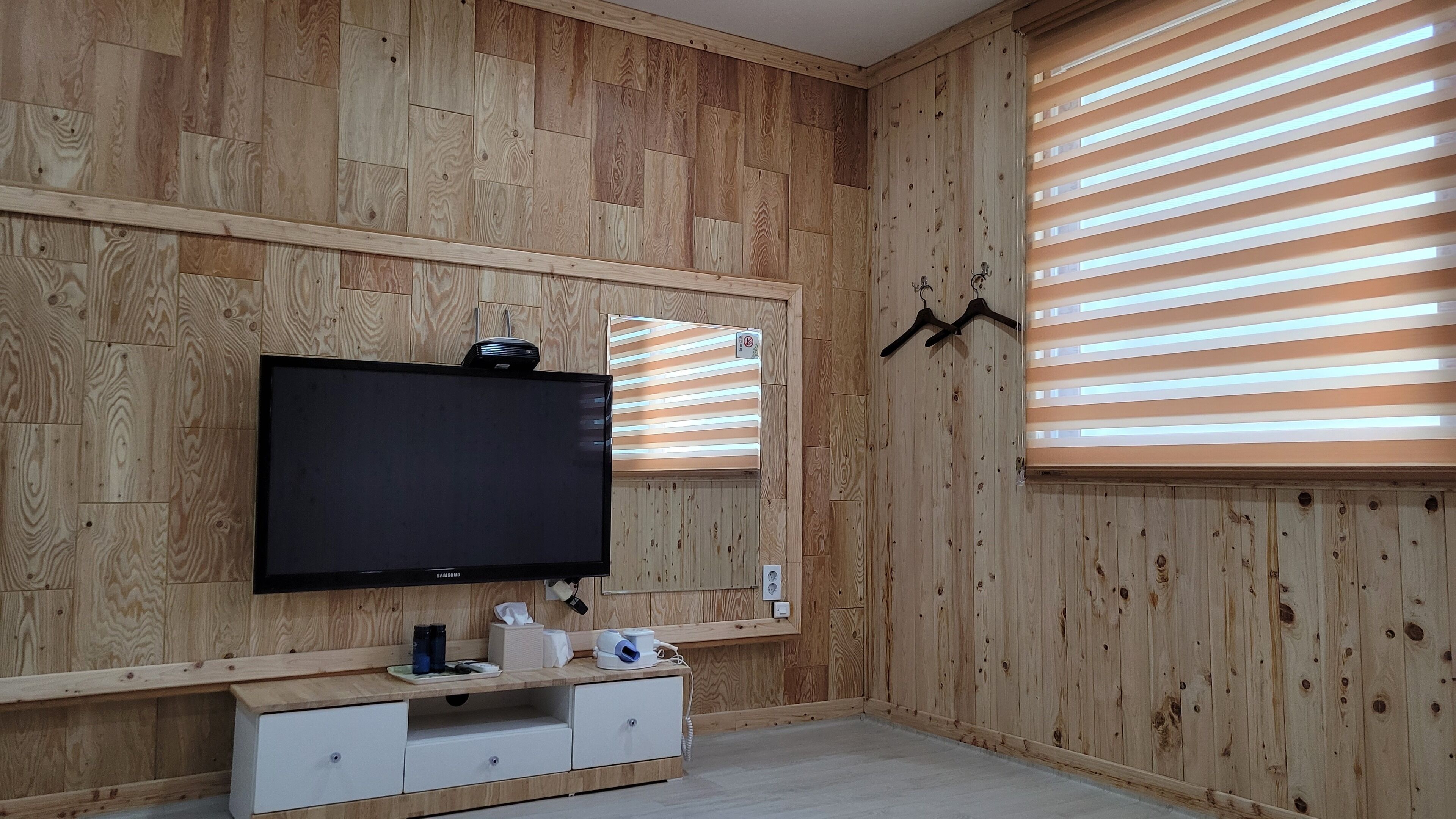 A room with wooden panel walls, a flat-screen TV mounted on the wall, a small white cabinet below, and a window with horizontal blinds.