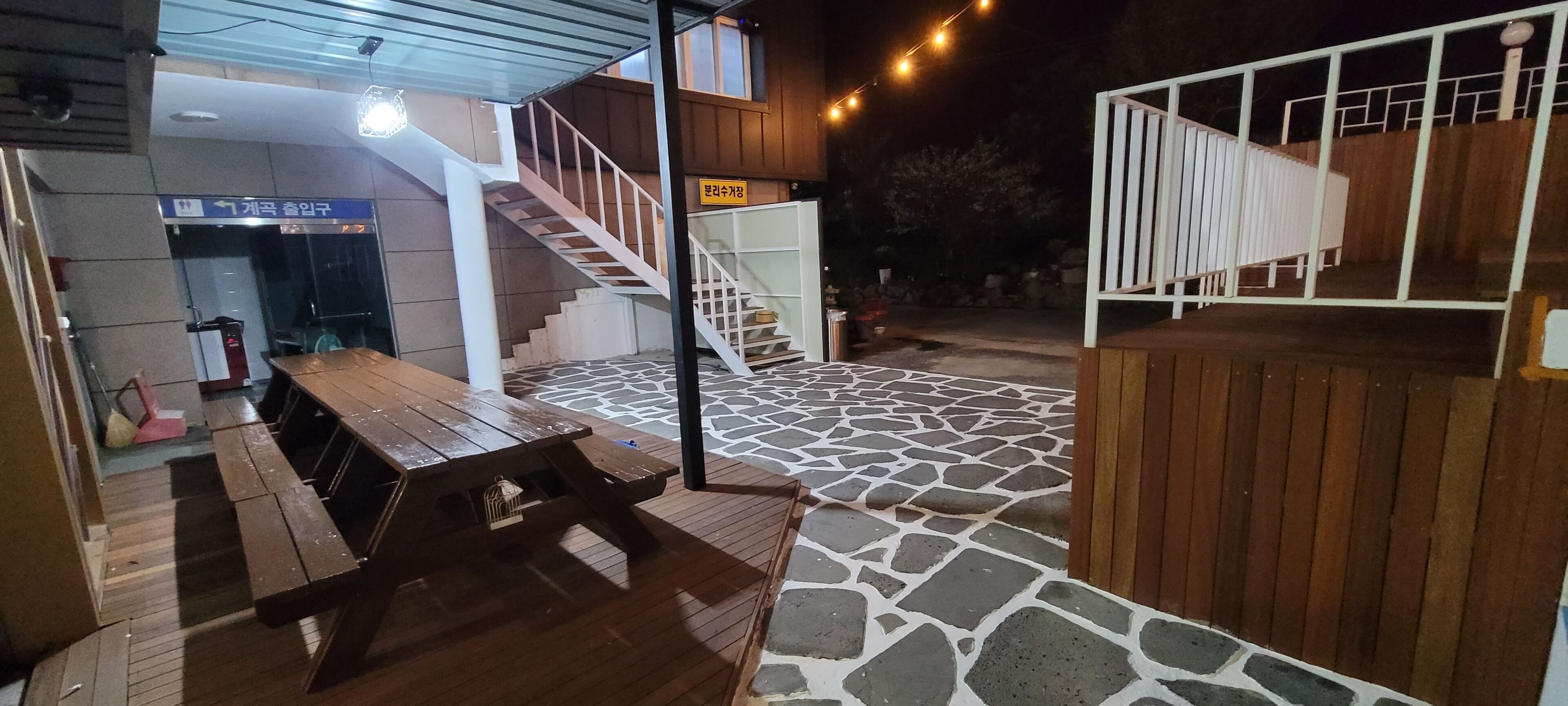 Ein Außenbereich mit einer Holzterrasse, einem Holztisch, Bänken, einer Treppe und einem steinbelagten Boden.