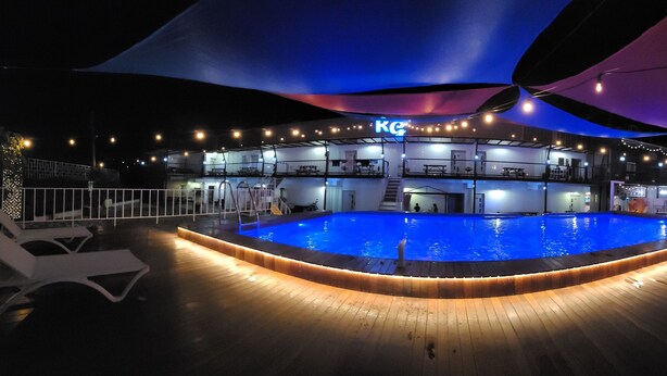 Ein Schwimmbecken mit Holzterrasse, Liegestühlen und einem überdachten Bereich mit einem 'KG'-Schild.