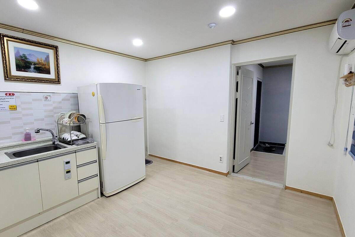 A compact kitchen with a refrigerator, sink, and a dish rack. A framed picture hangs above the sink, and a door leads to another room.