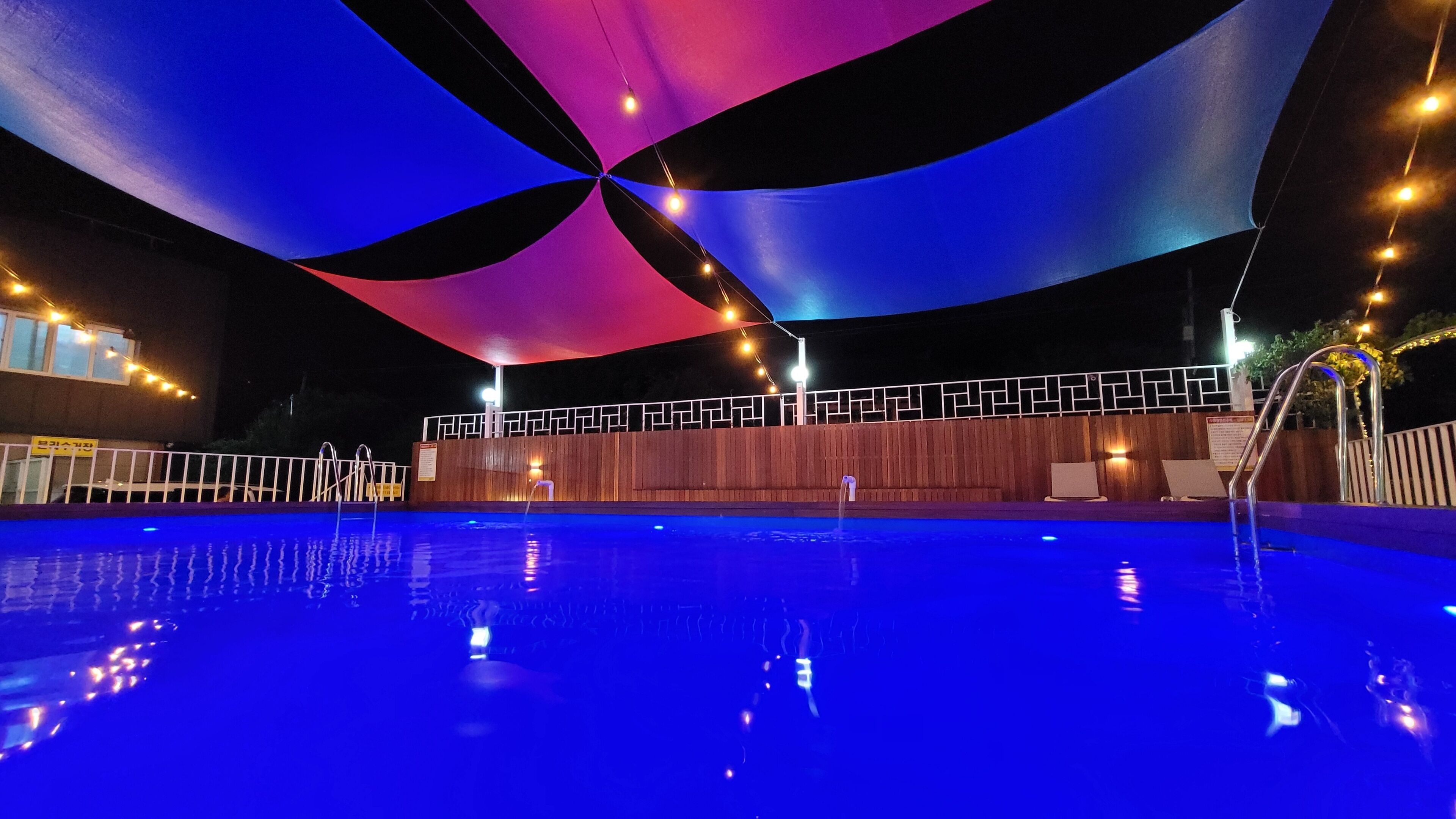 A swimming pool with a blue water surface, surrounded by a wooden deck and a covered area with a geometric pattern.
