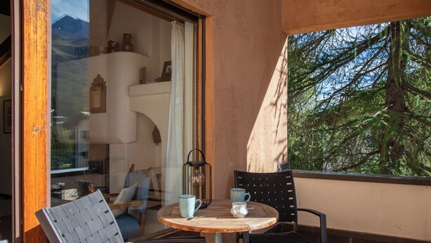A cozy outdoor seating area with a round table and two chairs, a lantern, and a view of a tree through a window.