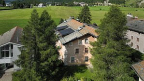 A residential area with a house featuring a sloped roof and solar panels, surrounded by lush greenery and mountains in the background.