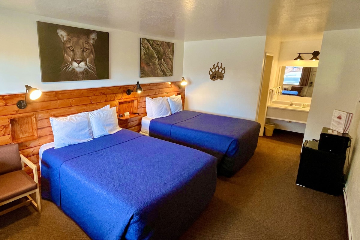 A hotel room with two beds, a wooden headboard, a nightstand, a chair, and a desk with a lamp.