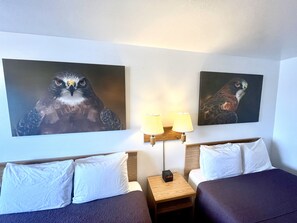 Habitación de hotel con dos camas, una mesita de noche con lámpara y dos cuadros de lechuzas en la pared.