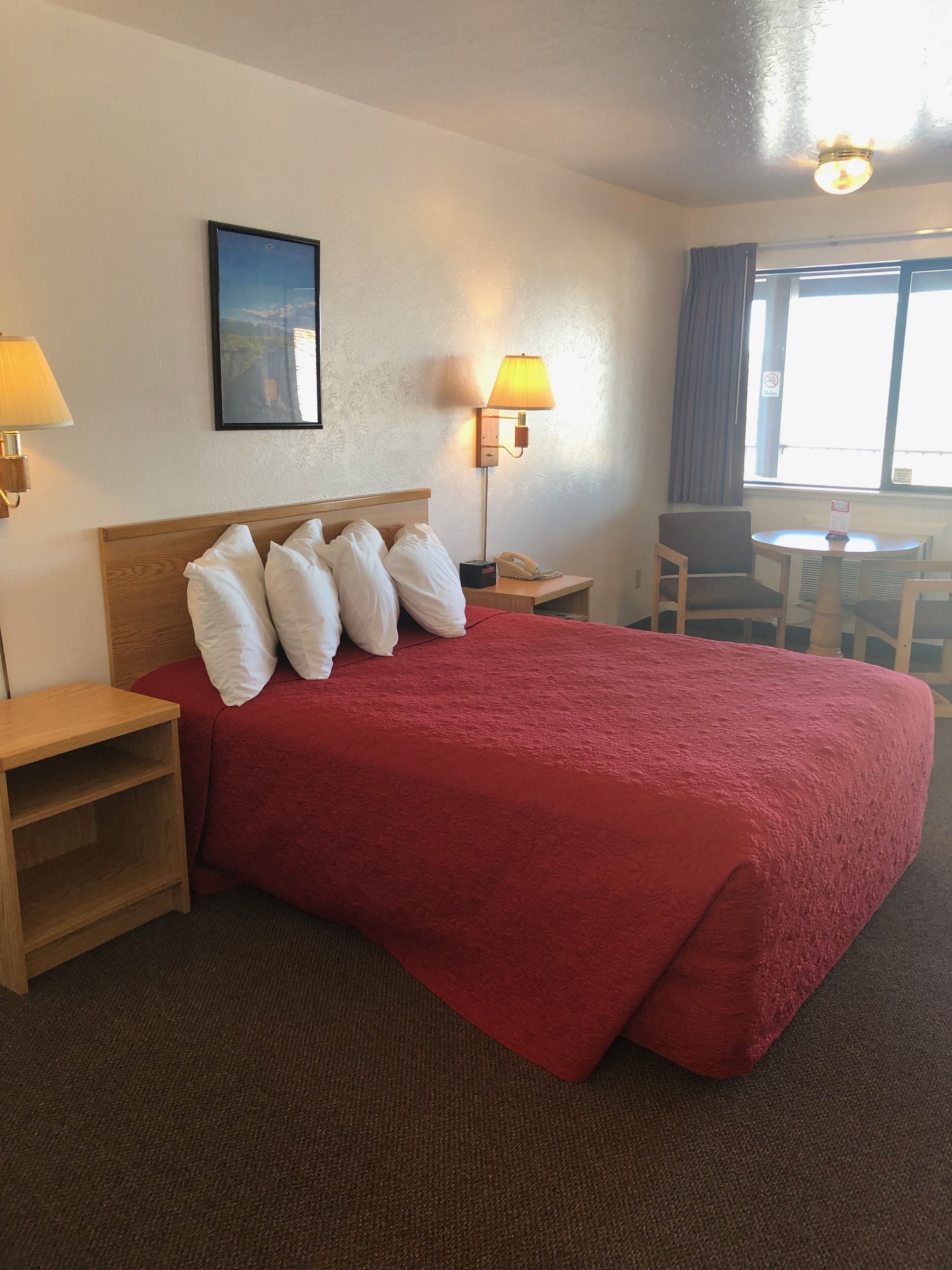 A hotel room with a large bed, two bedside tables, a desk with a chair, a window with curtains, and a wall-mounted lamp.