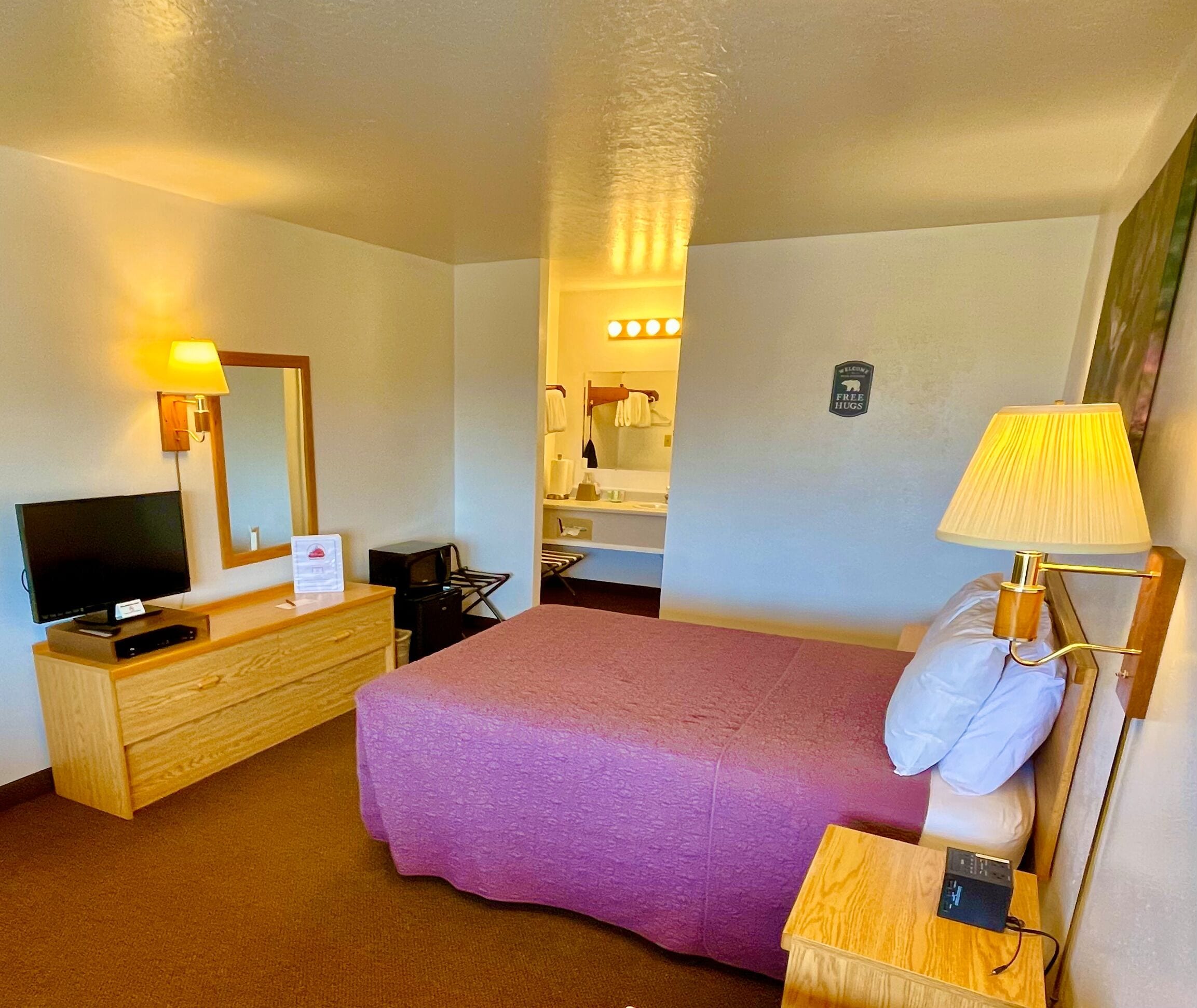 Habitación de hotel con cama, televisor, escritorio con lámpara, cómoda y baño visible a través de una puerta abierta.
