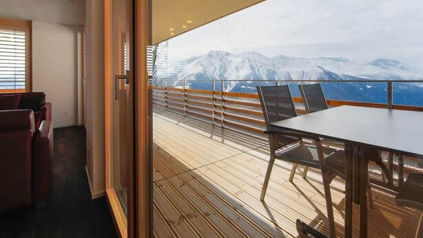 Ein Balkon mit Tisch und Stühlen, der Blick auf schneebedeckte Berge und ein Gewässer bietet.
