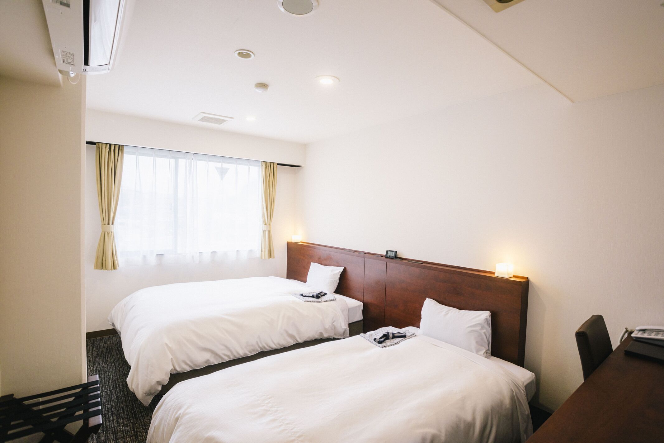 A hotel room with two beds, a wooden headboard, a desk with a telephone, and a window with curtains.