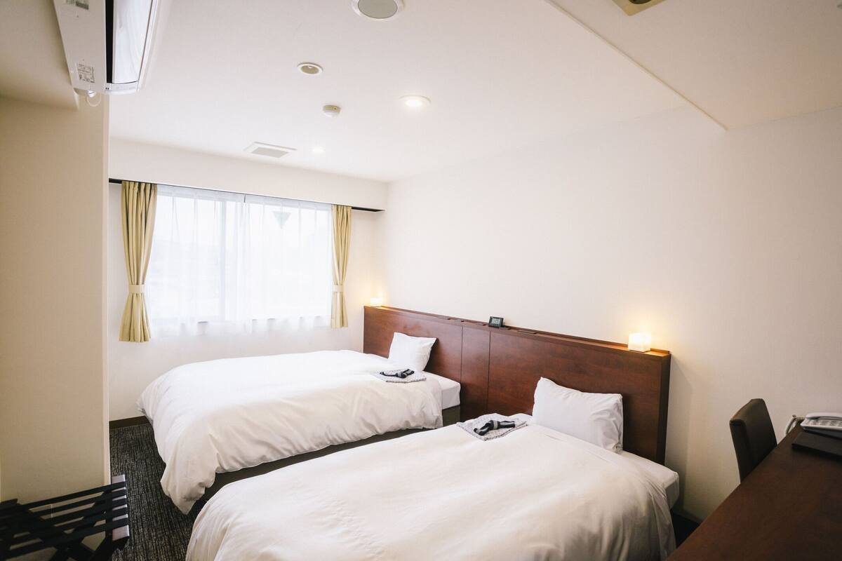 A hotel room with two beds, a wooden headboard, a desk with a telephone, and a window with curtains.