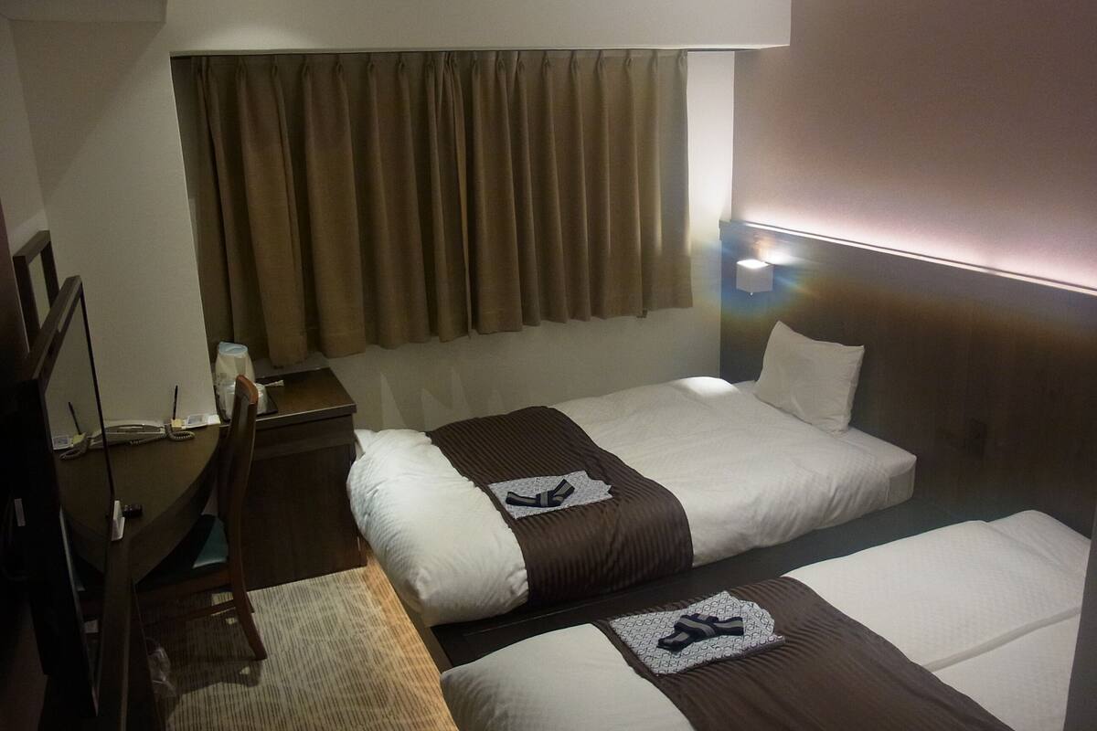 A hotel room with two beds, a desk, a chair, a lamp, and a window with curtains.