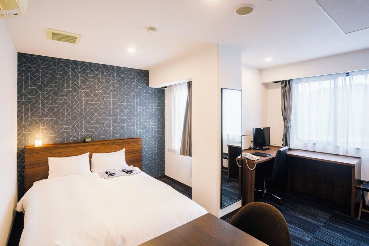 A hotel room with a bed, a desk, a chair, and a large window with curtains.