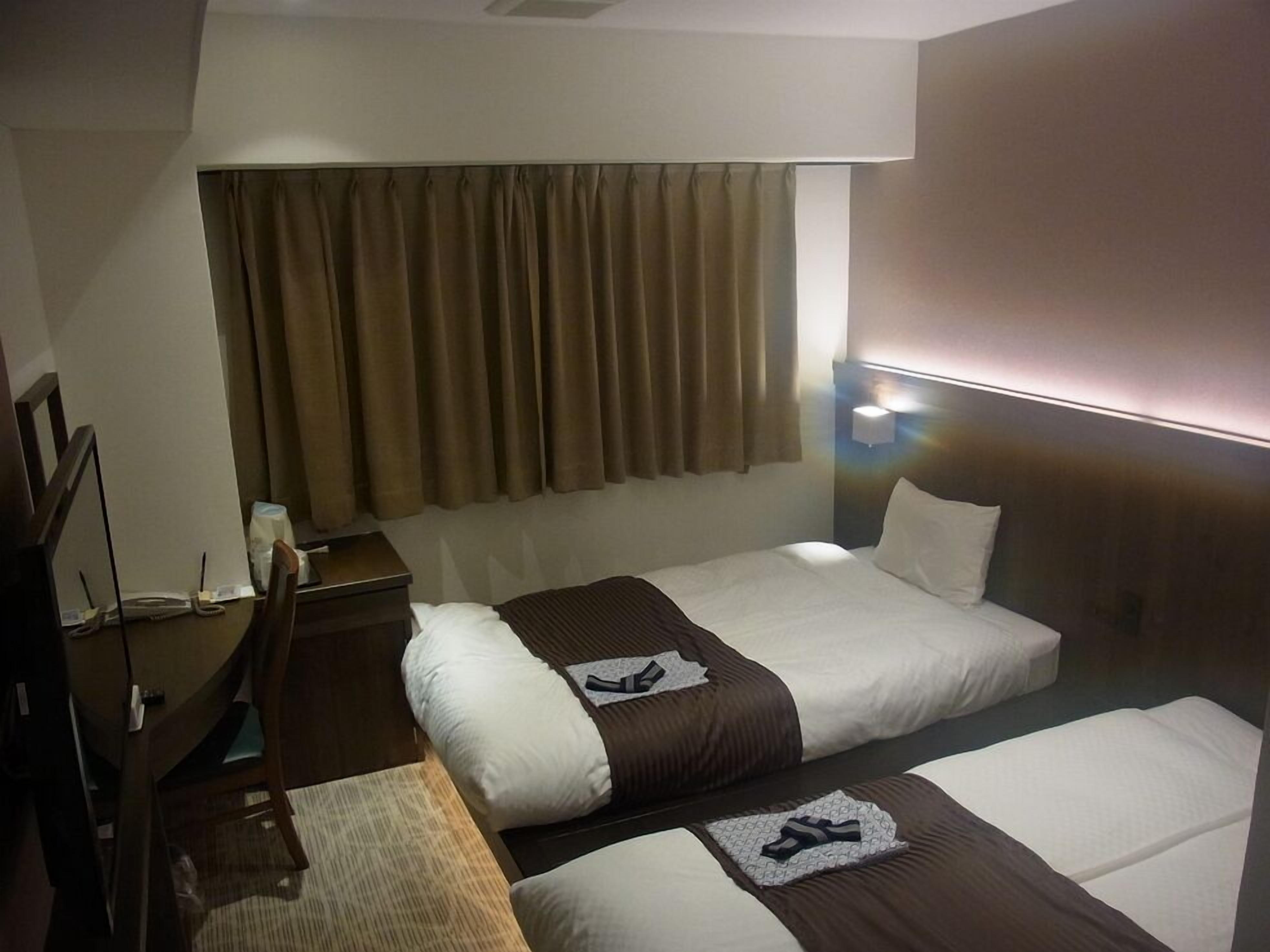 A hotel room with two beds, a desk, a chair, a lamp, and a window with curtains.