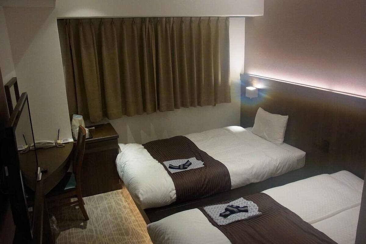 A hotel room with two beds, a desk, a chair, a lamp, and a window with curtains.