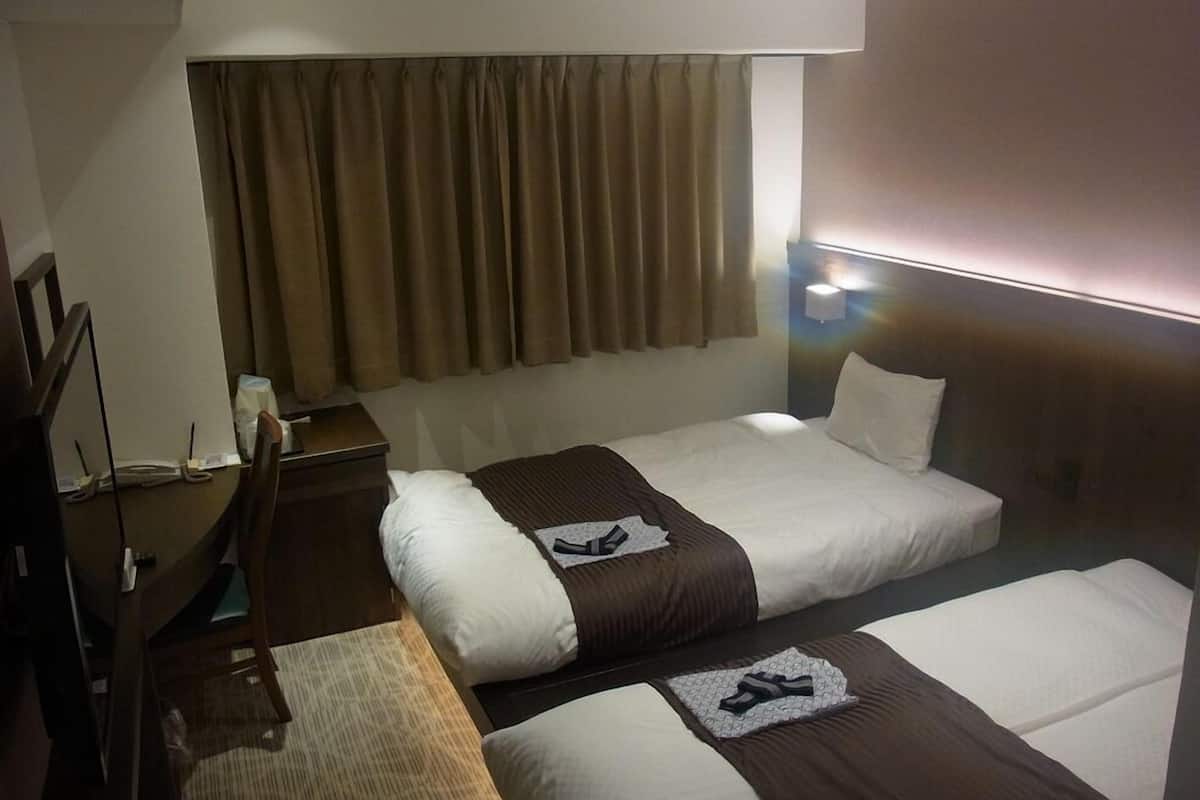 A hotel room with two beds, a desk, a chair, a lamp, and a window with curtains.