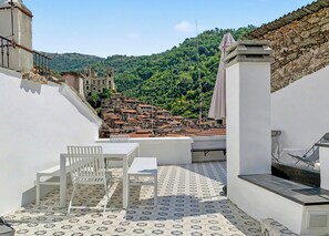 Um terraço no telhado com vista para uma aldeia no morro, móveis brancos e piso estampado.