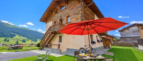 Eine Holzhütte mit Balkon, einem roten Sonnenschirm und einem Tisch mit Stühlen in einer grünen Wiese.