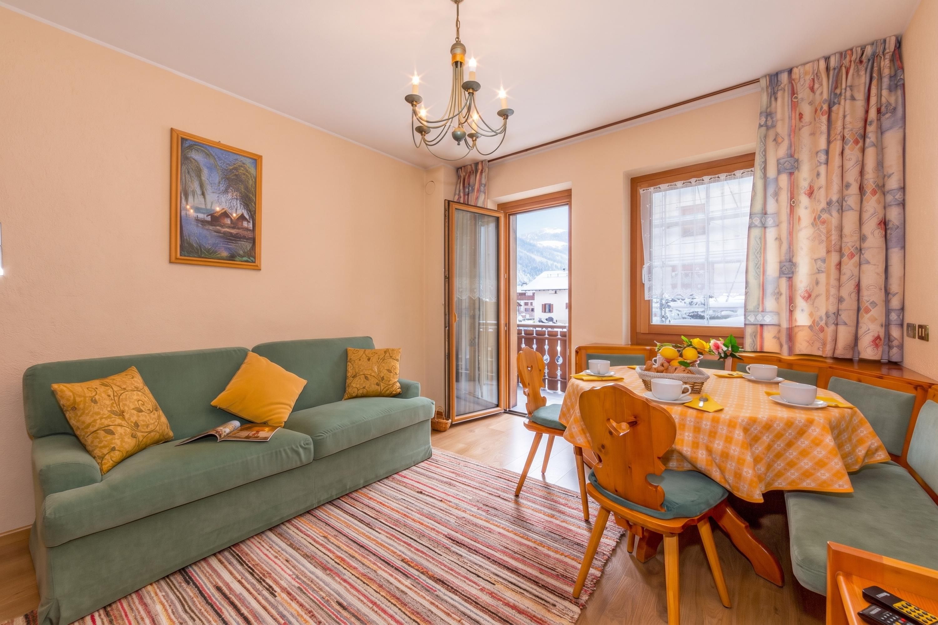 Una sala de estar acogedora con un sofá verde, una mesa de comedor con sillas, una araña y un cuadro en la pared.