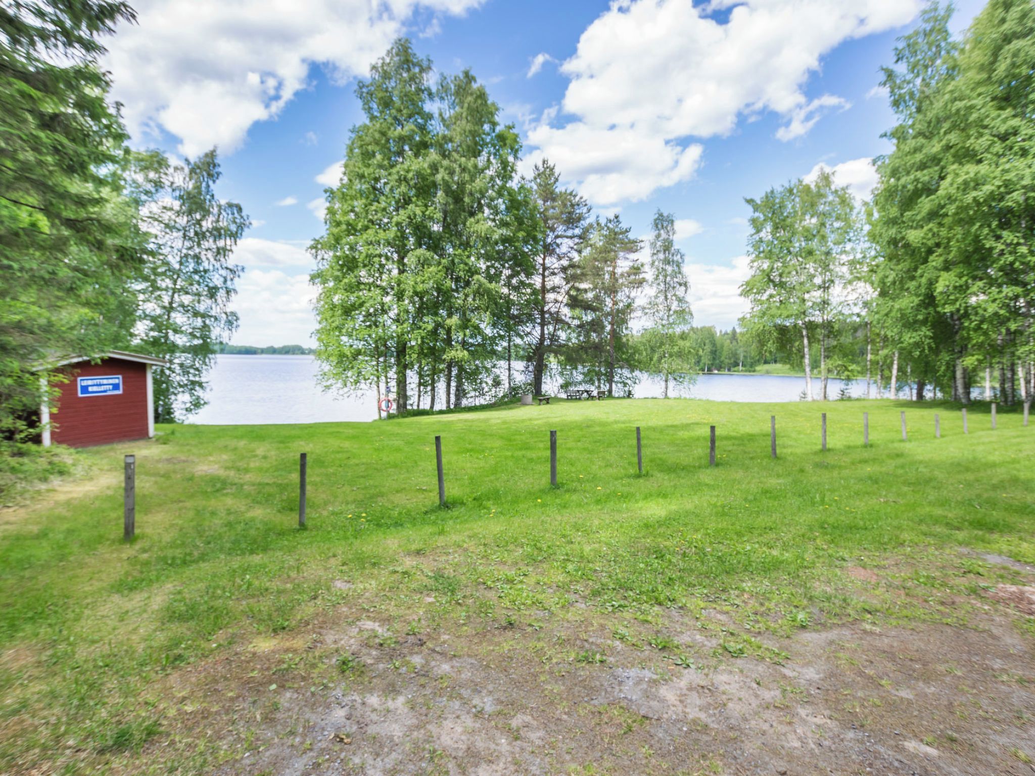 A grassy area with a fenced path leading to a body of water, a small red building, and trees.