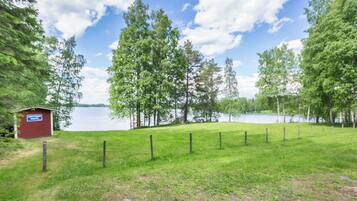 A grassy area with a fenced path leading to a body of water, a small red building, and trees.