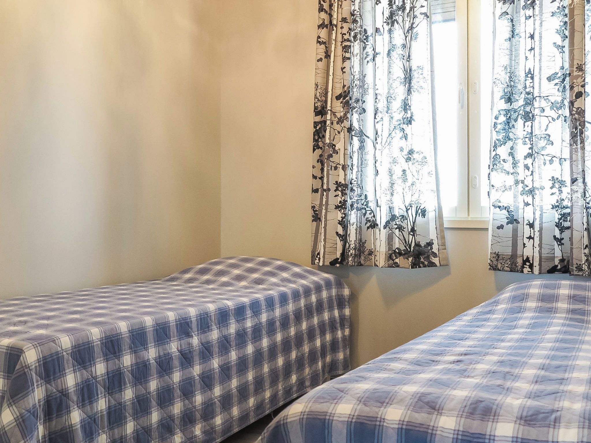 Una habitación con dos camas, cada una con una colcha a cuadros, una ventana con cortinas florales y una pared lisa.