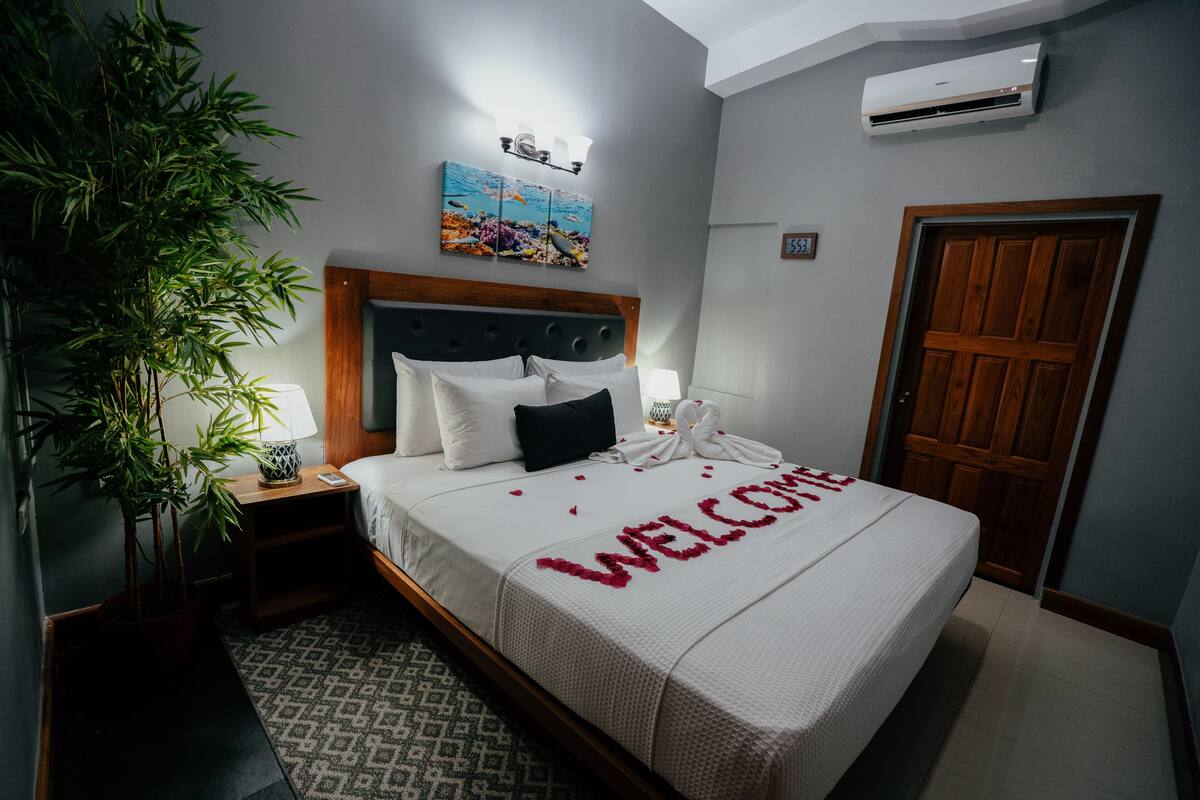 A neatly made bed with a welcome mat, a bedside lamp, a wall-mounted air conditioner, a wooden door, and a large potted plant.