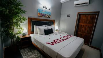 Una cama bien hecha con un tapete de bienvenida, una lámpara de noche, un aire acondicionado de pared, una puerta de madera y una planta grande en maceta.
