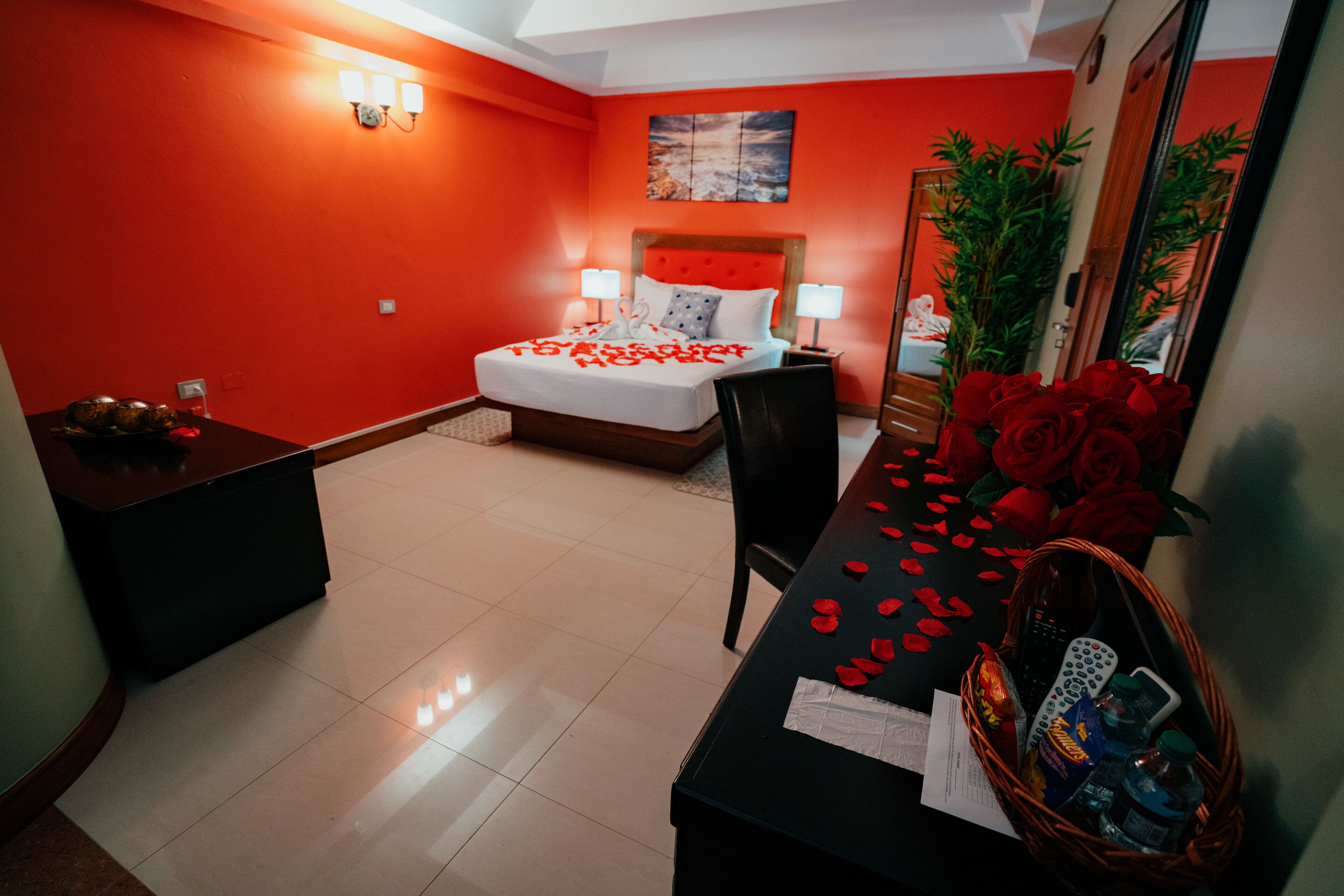 Una habitación de hotel con una pared roja, una cama con ropa blanca, una mesita de noche, un escritorio con una cesta de objetos y un espejo grande.