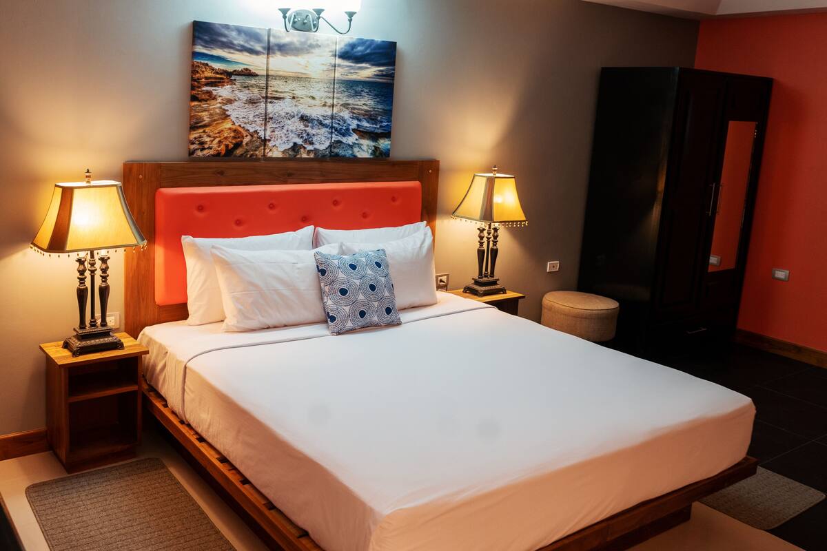 A neatly made bed with white linens, two bedside lamps, a headboard, and a painting above the bed.