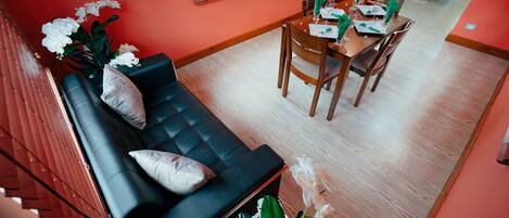 Une chambre d’hôtel moderne avec un canapé en cuir noir, une table à manger dressée pour deux personnes et une grande fenêtre équipée de stores.
